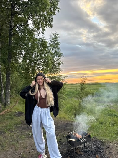 A young woman with long blonde hair stands in a clearing near trees, wearing a dark hooded jacket, a brown top, and light grey sweatpants. She is holding the hood of her jacket with both hands. In the foreground to her right, a metal grill with meat skewers is smoking. The background features a grassy field and a body of water under a cloudy sky with sunset colors. This scene is located in Bolshaya Izhora, Russia.