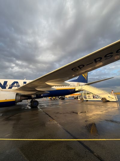 In Tillé, France, an airplane with the registration SP-RSS and "Ryanair" logo is parked on a wet tarmac under a cloudy sky. A mobile staircase is connected to the aircraft's entrance, and a person stands at the top of the stairs. The tarmac is wet, reflecting the surrounding light and aircraft. Other ground equipment, including a fuel truck, is visible in the background.
