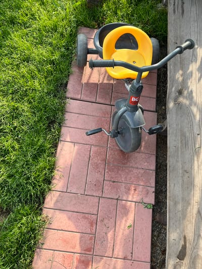 A yellow and gray child's tricycle is parked on a paved walkway next to a grassy area and a wooden plank. The tricycle has a yellow seat and handlebar grips, and a gray frame and wheels. The paved walkway is made of rectangular, reddish-pink bricks. The grassy area is lush and green, with some sunlight filtering through. The wooden plank is rough and weathered, with visible grain. The scene appears to be outdoors in a residential area.