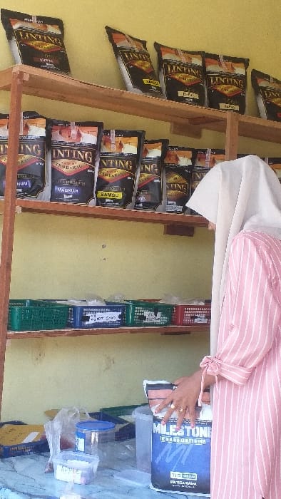 The image depicts an interior retail scene in Batang Kuis, Indonesia, characterized by a light yellow wall and natural illumination. A two-tiered wooden shelf unit occupies the mid-ground. The top shelf displays approximately seven black and gold pouches labeled "LINTING," indicating products such as rolling tobacco, with variants like "DUYUNG" and "VANILI" visible. The middle shelf features a similar arrangement of "LINTING" pouches, including "PREMIUM," "MADHUM," and "BAMBU" varieties. Below these pouches, multiple small plastic baskets—green, blue, and red—contain various packaged items, one blue basket having a label partially legible as "SEMU."
In the foreground, a white and grey marbled counter or table holds several clear plastic containers and bags. One rectangular container is labeled "APPLE," and a blue-lidded clear container is also present. On the right side of the frame, a person is visible from the chest upwards, wearing a long-sleeved pink and white striped garment with bow-tie cuffs and a light-colored hijab. Their right hand, adorned with two rings on the ring finger and pink nail polish, rests on a large black and blue bag labeled "MILESTONE TEELL," suggesting an interaction with the product.