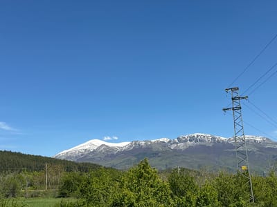 The image depicts a landscape near Manolovo, Bulgaria. A large mountain range with patches of snow is visible in the background. Below the mountain range, there's a green area with trees and vegetation. In the foreground, a steel electrical transmission tower and power lines are shown extending upwards into a vast blue sky.