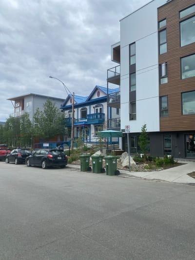 In Whitehorse, Canada, a street scene unfolds with a modern building dominating the right side, its facade a mix of brown wood paneling and white corrugated metal. A contemporary apartment building with balconies and large windows is visible on the left, behind a row of parked cars. Between these two structures, a colorful, older building with a bright blue roof and white trim stands out. The street is paved, with a sidewalk bordering the buildings. Several green garbage bins are placed along the sidewalk. A streetlight arches over the scene. The sky is overcast, suggesting a cloudy day. The overall impression is of a mixed-architecture urban environment.