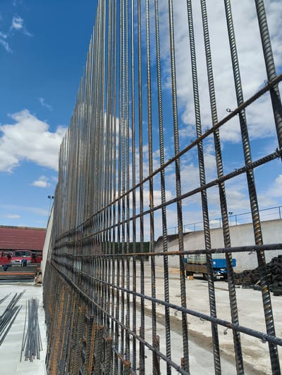 A construction site in Kladruby, Czechia, shows a large wall of vertical rebar, forming a cage-like structure. The rebar is metallic and appears to have some rust on its surface. Horizontal rebar crisscrosses the vertical bars, creating a grid pattern. In the background, a blue truck is visible, and a pile of tires is stacked to the right. To the left, a red vehicle with its doors open is parked near a building with a red roof. Scattered steel rods lie on the concrete ground in the foreground. The sky above is blue with scattered white clouds.