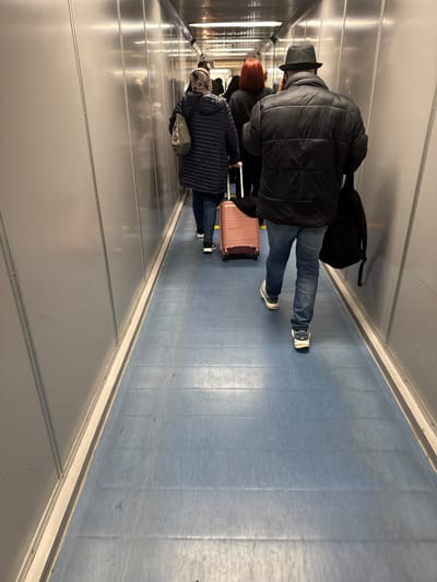 The scene depicts the interior of a jet bridge at Mohammed V International Airport (CMN) in Nouaceur, Morocco. A line of passengers is walking away from the camera through a narrow, enclosed corridor. The walls are constructed from light grey, reflective metal panels, arranged horizontally. The floor is covered with a textured, light blue material, showing distinct horizontal seams. Overhead lighting illuminates the space.

In the foreground, from behind, a male figure is visible, wearing a black puffer jacket, blue jeans, light-colored athletic shoes with dark accents, and a dark fedora-style hat. He carries a black backpack on his left shoulder. Immediately in front of him, a female figure, also viewed from behind, wears a dark blue quilted jacket, blue jeans, dark sneakers, and a light-colored, possibly patterned, headscarf. She is pulling a light pink hardshell rolling suitcase with an extended handle. Further down the corridor, behind the female, the back of a person with distinct red hair is visible, wearing dark clothing. Several other indistinct figures are present further in the distance, indicating a queue. All individuals are proceeding in the same direction.
