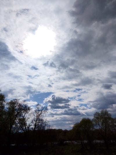 The image depicts a cloudy sky above a landscape, likely taken in the vicinity of Дерезовка, Russia. The sky is dominated by a mix of light and dark clouds, with a bright area suggesting the sun is obscured by the cloud cover. Below the clouds, the silhouetted forms of trees and vegetation are visible in the foreground. The scene suggests a day with unstable weather conditions.