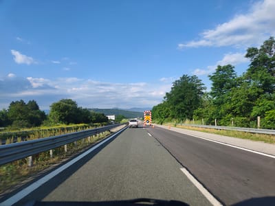 A first-person perspective, driving on a highway on a clear day, captures a scene with a bright blue sky dotted with white clouds. The highway stretches forward, marked with white lines. To the left, a silver guardrail runs alongside the road, bordered by green foliage and trees. On the right side of the highway, more trees form a dense green backdrop. In the distance, a white pickup truck is visible, followed by a large yellow and red traffic warning sign, indicating road construction or a hazard. Orange traffic cones are placed on the road ahead of the sign. Rolling hills and mountains are faintly visible in the background. The location is identified as the highway near Ravnishte, Bulgaria.