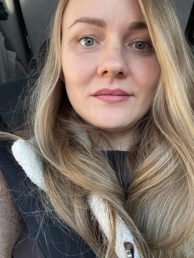 A close-up, eye-level portrait shows a light-skinned adult female inside a vehicle in Bolgury, Russia. She has long, wavy blonde hair with lighter highlights, parted on the left side and cascading over her shoulders and chest. Her eyes are blue-gray, and her light brown eyebrows are visible. Her lips are painted with a muted pink lipstick. Faint shadows are present beneath her eyes. She is wearing a dark-colored top, over which a cream or white fuzzy collar or trim is visible. A dark seatbelt strap crosses diagonally over her left shoulder and chest. A dark vehicle interior, including a dark seatback and what appears to be a headrest, is visible in the blurred background to the left. A textured brown fabric, possibly part of a coat or car seat, is visible on the far left edge of the frame. The subject is looking directly into the camera.