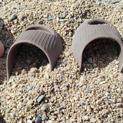 The image shows a close-up, eye-level view of two brown, pot-shaped objects partially embedded in a bed of small, light-colored gravel. The objects are curved and appear to be made of a ceramic or clay material, with vertical ribbed textures on their exterior surfaces. They are hollow and filled with smaller pieces of gravel and dirt. The gravel surrounding and within the objects is varied in size and color, with shades of tan, brown, grey, and white. The lighting suggests an outdoor environment under natural sunlight, casting subtle shadows. No people or identifiable city landmarks are visible. The scene is devoid of any context that would link it to a specific location in Torayghyrov, Kazakhstan.