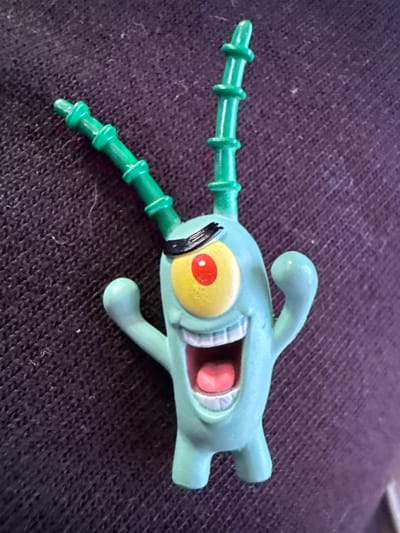 A light blue, plastic figurine of the cartoon character Plankton is positioned centrally on a dark purple, textured surface, likely fabric. The figurine depicts Plankton with his single yellow eye, a red pupil, and a black eyebrow. His mouth is wide open, revealing pink gums and white teeth, and his tongue is visible. Two green, segmented antennae extend upwards from his head. His arms are raised, and he has two small feet. The surface beneath the figurine has a ribbed or knitted texture, with visible threads and some minor lint. The lighting appears to be natural, casting subtle shadows. No specific location in Vyzyrka, Ukraine, can be identified from the image.