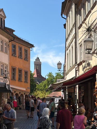 The image captures a vibrant street scene in Bamberg, Germany, likely during a sunny day. The architecture features traditional European buildings with colorful facades, particularly an orange building on the left adorned with windows and a whimsical stork figurine carrying a baby. On the right, a tall, cream-colored building with multiple arched windows and decorative elements looms. A street lamp with a star-shaped ornament is visible attached to this building. In the background, a prominent tower with a green spire, likely part of a historic church or cathedral, rises above lush green trees.
The street itself is paved with cobblestones and bustling with people. Shoppers browse stalls, some displaying clothing and other goods under awnings. Several individuals are walking along the street, engaged in conversation or looking at their phones. A man in a plaid shirt examines his phone in the foreground. A woman in a pink top and a woman in a white hijab are also visible among the crowd. The atmosphere appears lively and typical of a European market street.