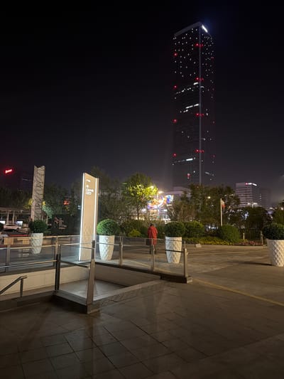 The image, taken at night, shows a cityscape in Jianye District, China. A tall skyscraper with lit windows is visible in the background. Closer to the foreground is a sign directing to ITC and other places. There's some greenery with small lights, and a person wearing a red shirt stands next to some modern, white outdoor furniture. The foreground features a pedestrian area with paving stones and metal and glass railings.