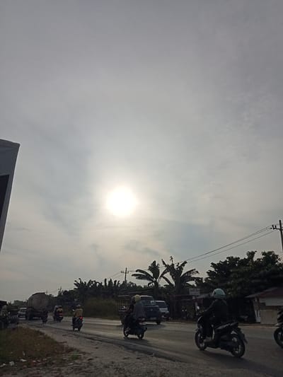 The image depicts a roadside scene in Kubang Jaya, Indonesia, under an overcast sky with the sun appearing as a bright, diffuse orb. On the left, the corner of a grey building is visible. The road is paved and shows signs of wear, with a dirt shoulder on the left. Several motorcycles and a few cars are present on the road. A large truck with a cylindrical tank is moving on the left lane. Three motorcycles are in motion, one with a rider wearing a yellow shirt and another with a rider in a grey jacket, both heading away from the camera. A third motorcycle, also moving away from the camera, has a rider wearing a blue helmet. A fourth motorcycle, on the right side of the road, is moving towards the camera, with its rider wearing a light blue helmet. Trees, including what appear to be banana trees, line the right side of the road, behind which are some small buildings. Utility poles and power lines are also visible against the sky. The overall atmosphere is that of a typical day in a developing urban or semi-urban area, with moderate traffic and ongoing activity.