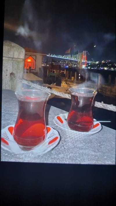 The image shows two tulip-shaped glasses filled with a dark red liquid, likely tea, on white and orange saucers. The glasses are placed on a textured surface, possibly a stone railing or table. In the background, a bridge illuminated with colorful lights spans across a body of water. There are also buildings with illuminated windows visible in the distance. A crescent moon is visible in the night sky. The overall scene depicts a nighttime view of a cityscape with a bridge as a prominent feature. The image has a grainy quality, suggesting it was taken in low light conditions. The specific location is not identifiable from the image itself, therefore the claim about Büyükçekmece, Turkey is not supported by visual evidence.