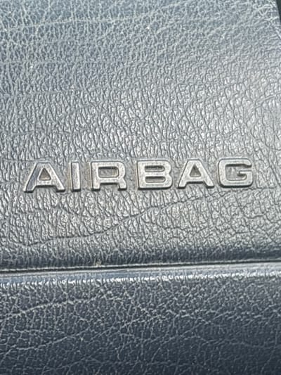 This media file is a close-up, vertically oriented photograph of the word "AIRBAG" embossed on a dark, textured surface, likely leather or a similar material. The letters are raised and metallic or silver-toned, standing out against the darker background. The texture of the material is finely grained, with a subtle, irregular pattern. The lighting creates shadows that emphasize the three-dimensionality of the embossed text. The image appears to be taken indoors, in a still environment, with no indication of time of day or weather. The focus is solely on the text and the material it is imprinted on, suggesting it might be part of a car interior component, such as a dashboard or steering wheel. There are no people or other objects visible in the frame. The text "AIRBAG" is clearly legible and is the sole subject of the image.