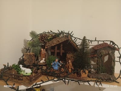 The image shows a Nativity scene display, likely set up for Christmas. It features small figurines representing Mary, Joseph, and other figures, set within a miniature landscape that includes a stable, a small structure resembling an archway, and a fenced enclosure with a sheep. The scene is decorated with pine branches, pinecones, and a string of green Christmas lights. The display is positioned on a shelf against a plain, light-colored wall. The overall impression is a homemade or traditional Christmas decoration.