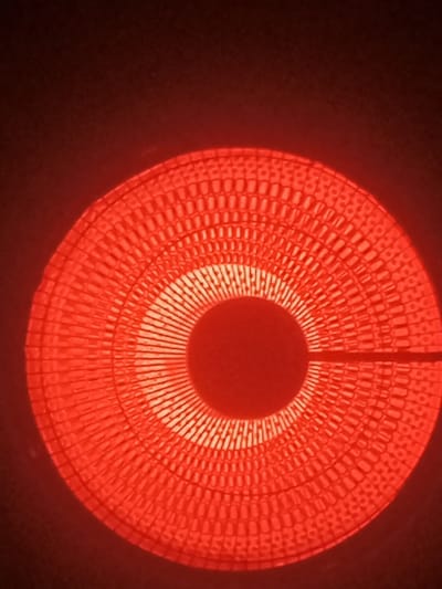 A red-hot electric stove burner glows intensely in the dark. The burner's circular design features concentric rings and a central void. A small, horizontal wire extends from the center. The overall impression is one of intense heat and a radiating, textured pattern.