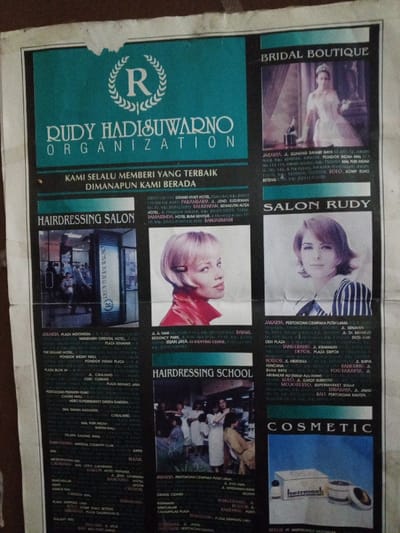 This image is a vertical photograph of a crumpled paper advertisement, affixed to an uneven, textured surface, likely a wall. The advertisement is for the "RUDY HADISUWARNO ORGANIZATION," a hair and beauty enterprise, with text primarily in Indonesian.
The top portion features a teal banner with a white logo displaying an 'R' within a laurel wreath, centered above "RUDY HADISUWARNO ORGANIZATION" and the slogan "KAMI SELALU MEMBERI YANG TERBAIK DIMANAPUN KAMI BERADA" (We always give the best wherever we are).
The page is divided into multiple sections:
- **Top Right, "BRIDAL BOUTIQUE":** Displays a woman in a white wedding gown holding a small bouquet, standing beside a dark curtain. Text below details business locations in Indonesian cities such as Jakarta and Solo.
- **Mid-Left, "HAIRDRESSING SALON":** Features an exterior shot of a modern salon with "RUDY HADISUWARNO" visible vertically on the glass door, and a headshot of a blonde woman with short, styled hair, wearing a red top. Text lists numerous salon locations across Jakarta and other Indonesian cities.
- **Mid-Right, "SALON RUDY":** Shows a headshot of a woman with medium-length brown hair, wearing a striped top and looking towards the viewer. Associated text lists further salon locations in Indonesian cities including Jakarta, Bandung, and Surabaya.
- **Bottom-Left, "HAIRDRESSING SCHOOL":** Presents an interior view of what appears to be a training session, with multiple individuals, primarily women, engaged in hairdressing activities. Location information is provided below.
- **Bottom-Right, "COSMETIC":** Contains a product shot of two "hairmask" containers. Further location details are listed beneath the image.
All explicit geographical references within the advertisement's text point to various cities and regions within Indonesia. The photograph itself captures this advertisement as it exists in a physical environment in the city of Keo Kouang, Laos.