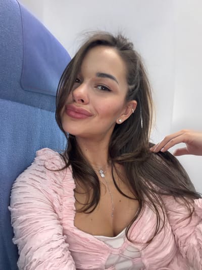 A young woman with brown hair is reclining on a blue surface, possibly a sofa or lounge. She is wearing a pink, textured long-sleeved top and a silver necklace with a butterfly or floral pendant. A hand is visible in the upper center of the image, gently touching her hair. The woman has fair skin, defined eyebrows, and is wearing lipstick. She has a small stud earring in her ear, and a piercing further up the ear. The background is out of focus and appears to be a plain, light-colored wall. No information about Shunyi District, China is discernible from this image.
