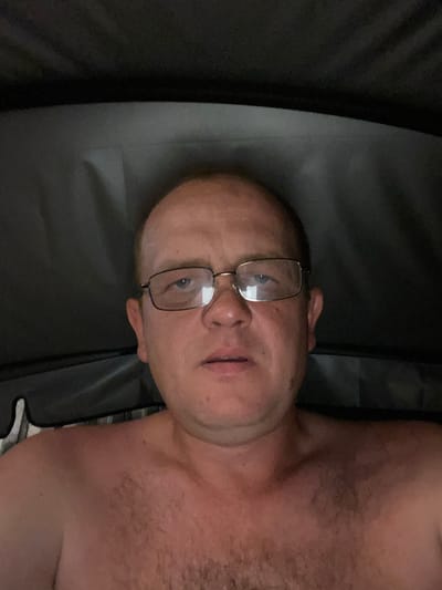 The image shows a close-up, low-angle, upside-down shot of a fair-skinned adult male with a bald head and glasses. He is lying down, and the background appears to be a dark, possibly outdoor setting with textured surfaces. The lighting is dim, suggesting it might be evening or nighttime. The man's face is partially visible, with his eyes open and looking towards the camera. He has a neutral expression. The visible portion of his body appears to be his chest and shoulder, which have some hair. There is no discernible text or specific activity taking place. The location context provided is Әуезов ауылдық округі, Kazakhstan, but there are no visual cues in the image to confirm this.