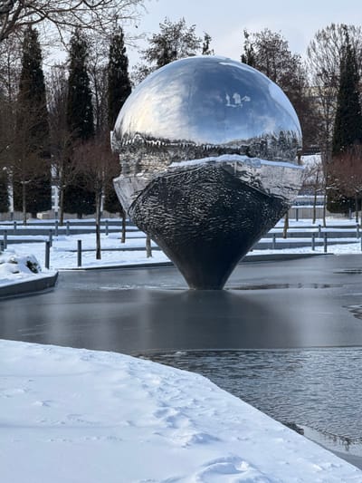 The image depicts a winter scene featuring a large, spherical art installation situated within a partially frozen body of water in a landscaped urban park in the city of Plodorodny, Russia.
The central object is a prominent sculpture, roughly 5-6 meters tall, composed of two distinct halves. The upper hemisphere is highly polished, reflective metal, possibly chrome, which mirrors the pale, overcast sky and the silhouettes of surrounding trees. A noticeable layer of white snow and numerous icicles are present along the sculpture's equator, marking the transition point between the two halves. The lower half of the sculpture is a dark, textured, conical form, appearing to emerge from or be supported by the water.
In the foreground and surrounding the sculpture is a body of water, partially covered with dark, reflective ice patches, while some areas of open water show slight ripples. Snow blankets the banks adjacent to the water and extends across the ground surfaces in the background. The background features a mix of bare deciduous trees and tall, dark evergreen trees, likely cypress or thuja, indicating a cold climate. Paved pathways or pedestrian areas, also covered in snow, are visible further back, interspersed with dark, short bollard-like structures. No individuals are present in the scene, and no specific actions or interactions are depicted.