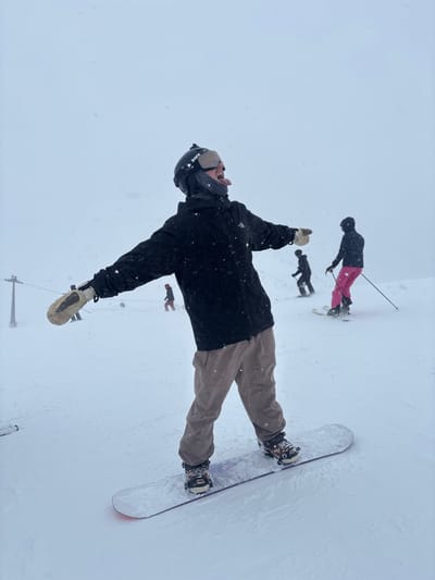 The image depicts a snowy ski slope in St. Christoph, Austria, under an overcast sky with active snowfall. In the foreground, a person is standing on a white snowboard with bindings, facing slightly left of the camera. The individual wears a black jacket with a visible "The North Face" logo, tan/brown snow pants, light-colored mittens, and a black Bern brand helmet with goggles and an attached GoPro camera. Their head is tilted backward with their tongue extended, and both arms are outstretched to the sides. In the midground and background, several other individuals are visible on the slope, engaged in skiing or snowboarding. One skier on the right wears a dark jacket and bright pink pants, holding ski poles. A portion of a metal ski lift support structure is visible on the far left. The entire scene is covered in white snow, with numerous snowflakes visible in the air.