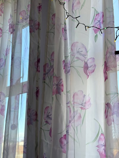 Close-up shot of pleated floral curtains in front of a window with a string of lights hanging to the left. The curtains are light-colored with a pattern of purple and pink tulip-like flowers with green stems. The pleated design creates horizontal layers. Glimpses of daylight are visible through the gaps in the curtains. The string lights have a black wire and small multi-colored LED bulbs. The scene appears to be indoors. It is not possible to provide a precise location within Craigavon, United Kingdom, as no specific landmarks or indicators are present in the image.