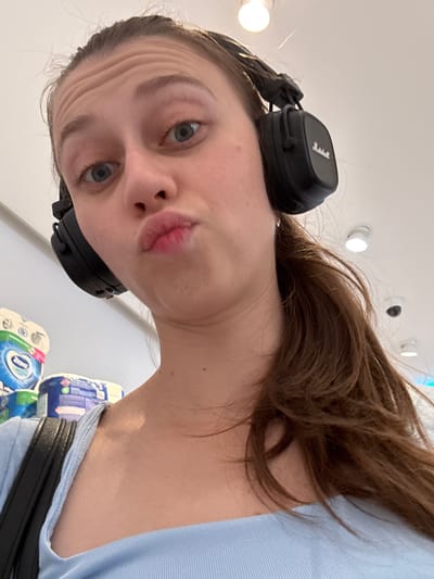 The image shows a young woman with brown hair pulled back in a ponytail, wearing black over-ear headphones. She is looking at the camera with her lips pursed in a duck face pose. The woman is wearing a light blue ribbed top and a black strap is visible over her shoulder, suggesting she is wearing a bag. In the background, there are stacks of packaged goods, possibly hygiene products like toilet paper or diapers, suggesting a retail environment such as a supermarket. The lighting is bright, with ceiling lights visible. The overall impression is a casual, selfie-style photograph. There is no indication of the location being Erlenbach am Main, Germany.