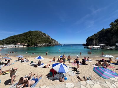 The image displays a panoramic view of a sandy beach situated within a clear, turquoise bay in Palaiokastritsa, Greece, under a vivid blue sky with scattered wispy clouds and a high-altitude contrail. Steep, densely vegetated hills and rocky outcrops flank both sides of the bay. On the left side of the bay, buildings with light-colored walls and reddish roofs are visible at the base of the hill. On the right, a small dock or pier extends into the water, with several white motorboats moored alongside. The immediate foreground consists of a flat surface paved with irregular light-colored stone tiles.

Numerous individuals are present on the beach and in the water. On the light-tan sand, people are seen sunbathing on towels, sitting or reclining under various beach umbrellas (including striped blue and white, red and blue, and a multi-colored pink, blue, and green umbrella). In the foreground, two men lie prone on striped towels. One man, in black shorts, lies on a blue and white striped towel; the other, in blue shorts, lies on a similar towel. Near them, a woman in light attire is bent over. Other groups are seated or standing on the sand. In the bay, many people are swimming or wading in the clear, shallow water, while a few are further out. Yellow buoys are visible marking an area in the water. Scattered personal items, such as bags and shoes, are visible near the umbrellas.