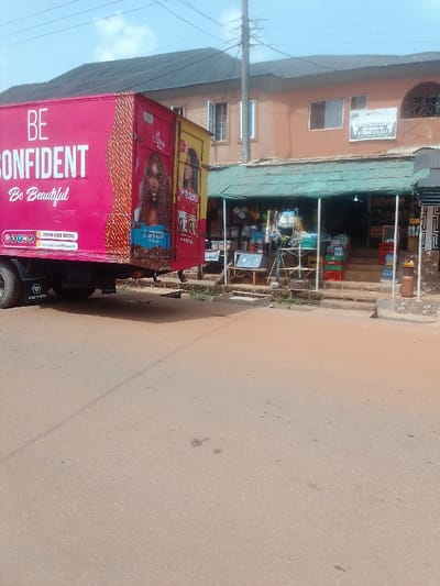 The image shows a pink truck parked in front of a shop in Auchi, Nigeria. The truck has the words "BE CONFIDENT Be Beautiful" printed on the side. The shop is a small, open-air structure with a green awning and a variety of goods on display. The shop is located in a residential area with a two-story building behind it. The street in front of the shop is paved and appears to be dusty.