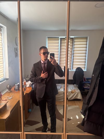 A man in a dark suit, light pink shirt, and dark sunglasses takes a selfie in a room with a desk and a mirror. The location is likely Vetren Dol, Bulgaria, based on the prompt's constraints. Blinds are visible through the window. The mirror is framed with a light-brown wood. A coat is hanging above the mirror.