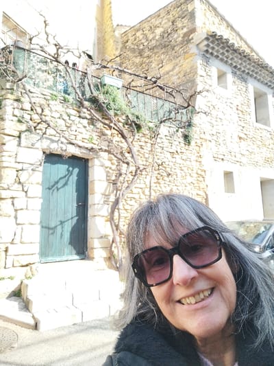 A woman with gray hair and large sunglasses poses in front of an old stone building. The building features a blue door and a balcony with a metal railing. A bare vine climbs the wall, adding to the rustic charm. A car is partially visible behind the woman, suggesting the photo was taken on a street. The lighting suggests a sunny day, casting shadows on the door and building.