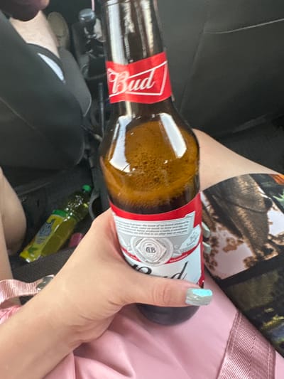 The image shows a close-up of a person's hand holding a brown glass bottle of Budweiser beer. The beer bottle has a red label with the Budweiser logo. The hand has light-colored nail polish with a blue design on one fingernail. The person is wearing a pink garment. In the background, a green bottle and part of a black car interior are visible. The scene appears to be inside a vehicle. There is no information within the image to suggest a specific location in the city of Ленин, Kyrgyzstan.