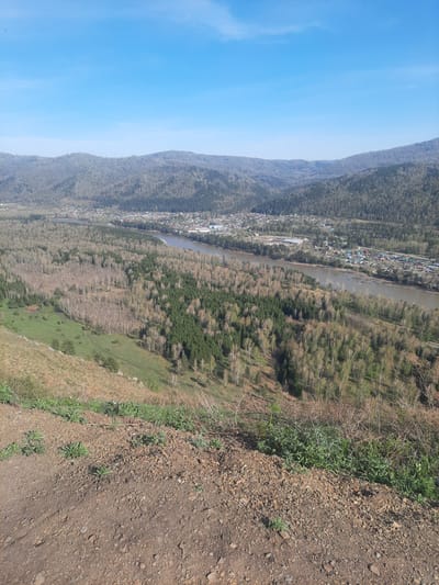 The image presents a landscape view from an elevated position, likely a hillside or mountain, overlooking a portion of the city of Aya in Russia, as suggested by the context. The right foreground is dominated by dirt and rocks, transitioning into sparse vegetation. Beyond this, a slope leads down to a mix of trees and grass. A river winds through the scene, flanked by bare trees and some evergreen forest sections. On the left side of the river, the city of Aya is visible, nestled against a mountain range which extends into the left side of the frame. Above the mountains is a clear blue sky. The overall composition suggests an autumn or early spring setting, given the mixture of bare and green foliage. The scene lacks any human presence.