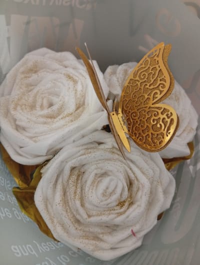 A bouquet of three white, rose-shaped decorations, possibly made of fabric or cotton, are arranged together. Each rose is adorned with scattered gold glitter. A golden butterfly decoration, with intricate filigree patterns, is perched on one of the roses, its wings spread. The bouquet is partially wrapped in gold material. The background is blurred and appears to be a light-colored surface with faint, translucent text or patterns. The overall scene suggests a decorative item, possibly for a gift or event. There is no identifiable location in the city of Seýdi, Turkmenistan, visible in the image.
