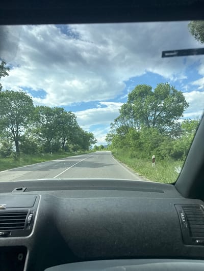 The image shows a view from inside a car driving on a road. On either side of the road are trees and greenery. The sky is visible through the car window, showing clouds and blue sky. The dashboard of the car is visible in the foreground. The location appears to be in a rural area, possibly near Studeno Buche, Bulgaria, given the landscape and road style. There are no people visible in the frame. A road marker pole is visible.