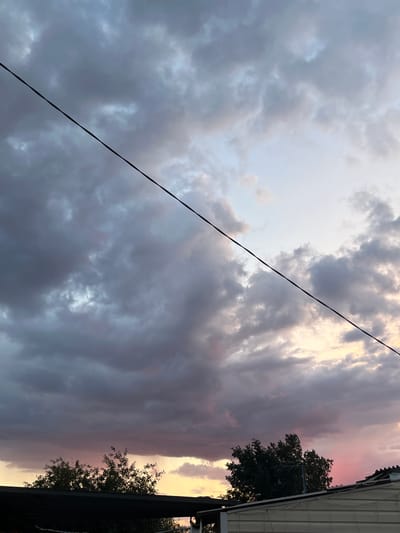 The image depicts a sky filled with dramatic, dark gray and purple clouds, tinged with soft pink and yellow hues near the horizon. A thin, black wire stretches diagonally across the frame, dividing the sky. In the lower portion of the image, the tops of dark green trees are visible against the colorful sunset sky. A section of a light-colored building with a dark-colored awning is also present at the bottom, partially obscuring the trees and sky. The scene appears to be an outdoor setting, possibly a residential area, at dusk. The location in Rakhinka, Russia, is not visually identifiable from the image itself.