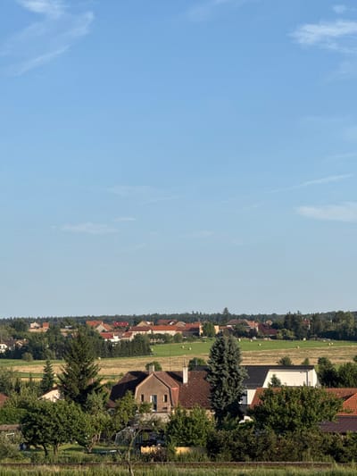 The image captures a rural landscape in Nučice, Czechia, under a clear blue sky with scattered wispy clouds. The foreground features dense greenery and trees, with hints of a wooden fence or railway tracks running horizontally across the bottom. In the mid-ground, several houses with reddish-brown roofs are nestled among trees. One prominent house in the center has a light brown facade and a chimney. Further back, a village stretches across the horizon, characterized by a cluster of buildings with various colored roofs, interspersed with fields and trees. Several round bales of hay are visible in the fields. The scene depicts a peaceful, sunlit day in a village setting.