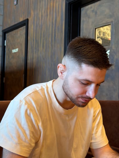 The scene depicts a male individual seated within an indoor establishment located in Bretton, United Kingdom. The subject, an adult male with light skin, dark brown hair styled in a high fade, and a short beard, is positioned facing slightly to the right of the frame with his head tilted downwards, his gaze directed below the visible area. He wears a plain, light yellow, short-sleeved crew-neck t-shirt. The background features a wall with a distressed, vertically striped texture in tones of brown, gold, and dark grey. Two dark-framed doors are visible. The door on the left bears the word "TOILET" written vertically in light lettering. The door on the right includes a small, square window with patterned or frosted glass, behind which blurred circular light sources are discernible. A segment of a brown, upholstered seating surface, possibly a booth, is present on the right side of the image. No explicit action or interaction is occurring beyond the subject's seated posture and downward focus.