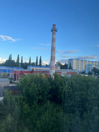 The media file captures a view of Leninogorsk, Russia, with a large smokestack dominating the left side of the frame. The smokestack is red and white striped and has a metal platform at its top. Behind the smokestack, there are several brightly colored industrial buildings with red and blue roofs. Further in the background, a row of tall, multi-story residential buildings with yellow and blue facades are visible. Dense green trees line the foreground and sides of the industrial buildings, obscuring some of the structures. The sky is a clear, bright blue with a few wispy white clouds. The lighting suggests it is daytime, likely late afternoon given the angle of the light and the overall clarity of the scene. There are no people visibly active in the image, and no text is discernible. The scene depicts an industrial and residential area, possibly on the outskirts of the city, under a pleasant sky.