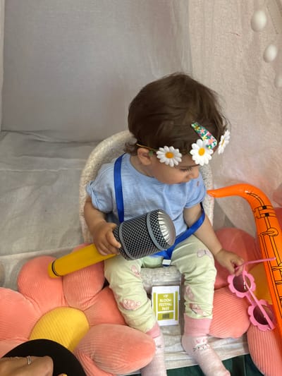 In the image, a baby is lying down, holding a toy microphone. The baby is wearing a light blue t-shirt, light green pants with a butterfly pattern, and pink socks. A floral hairband with white daisies is on the baby's head. Behind the baby, there are large, soft, flower-shaped cushions in pink and yellow, and an inflatable orange object, possibly a toy. A "VIP BLOOM FESTIVAL PASS" is visible on the baby's pants. The scene appears to be indoors, possibly in a playroom or at a festival, given the pass. The lighting suggests daytime. The overall mood seems playful and cheerful.