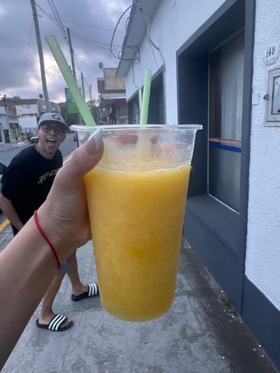 A hand holds a large cup of yellow slushie with two green straws. Behind the drink, a man wearing a hat and glasses playfully sticks his tongue out. The background shows a street scene with buildings and overcast skies. The photo is taken at an angle. The person holding the drink is wearing a red string bracelet and the legs of a person in sandals are visible.