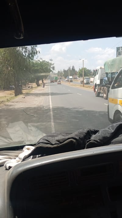 A view from inside a vehicle looking out onto a sunny street. The road has a white dividing line and cars and trucks travel in both directions. On the left, a dirt shoulder is lined with trees. Overhead, the sky is partially cloudy. Part of the vehicle's dashboard and windshield are visible in the foreground. The windshield has a small crack in it. A dark garment is draped on top of the dashboard with a white cloth underneath it.
