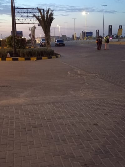 The image depicts an outdoor scene in Sidi Abdel Rahman, Egypt, captured at twilight. The viewpoint is from a low angle, looking out across a street. The street itself is divided into three distinct surface types: a dark brick-paved walkway on the right, a paved section in the middle, and a raised curb marked with alternating yellow and black stripes on the left.
Several people are visible further down the street, near a set of street lights. The environment includes several tall buildings, various road signs, shrubs, and a palm tree. A black car is visible parked by the left side of the street.