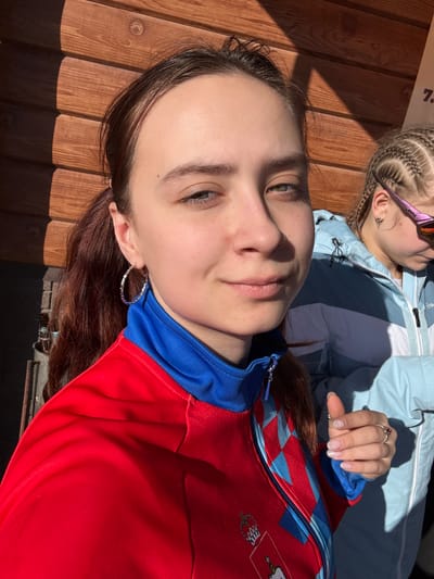 The image shows two young women from a low-angle view. One is wearing a red jacket with a blue collar and a pattern on the shoulder, while the other wears a light blue jacket. The first woman has brown hair and is wearing hoop earrings. The second woman has braided blonde hair and is wearing sunglasses. The background appears to be a wooden structure. A partial sign in the top-left corner suggests this might be a public place or venue. Based on the clothing and apparent location details, it can be surmised that the picture was taken in Gory, Russia.