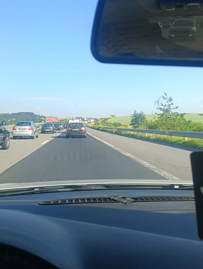 The image captures a view from inside a vehicle, looking forward and slightly to the right, on a highway likely near Vakarel, Bulgaria, based on visual cues such as the style of road signs and surrounding landscape. The sky is clear blue, indicating sunny weather. The highway is multi-lane with traffic in the distance, suggesting a moderate to heavy flow of vehicles. Several cars are visible on the road, including a silver Chevrolet sedan with license plate "PA 0471KK", a black BMW wagon with license plate "W 9223BT", and other vehicles further ahead. The road is paved with asphalt and has white lane markings. A metal guardrail runs along the right side of the highway, separating it from a grassy verge and then rolling hills covered with green fields and some trees. Billboards are visible in the distance on the left side of the highway, one of which appears to be red and white, with text that is not fully legible but might be an advertisement. Another yellow billboard with the text "erbet" is also visible further down the road. The scene depicts a typical highway environment with vehicles in transit under a clear sky.