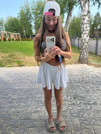 A young woman with long, brown hair, wearing a white baseball cap with a pink band and a zebra-print bikini top, holds a smartphone in her right hand, taking a selfie. She is also holding a colorful object, possibly a toothbrush, in her left hand near her face. She is wearing a short, white skirt. The woman is standing on a paved walkway made of grey, rectangular stones. To her left, there is a grassy area with trees, including a prominent birch tree with white bark. In the background, a black metal fence and some wooden structures can be seen. The lighting suggests it is daytime with bright sunlight, casting shadows on the walkway. The overall mood appears cheerful and relaxed. There is no visible text in the image. The context of Morozovo, Russia, is provided as external information.
