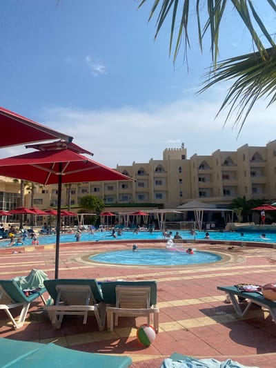 The image shows a hotel swimming pool area in Monastir, Tunisia. The pool is lined with red and white striped tiling and surrounded by lounge chairs with red umbrellas providing shade. People are swimming and playing in the pool, including some playing water polo with a net. A multi-story hotel building is visible in the background. A palm tree is visible in the upper left corner.