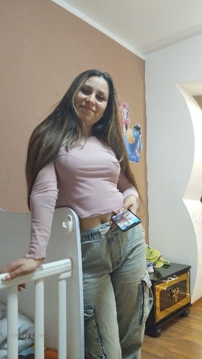 The image depicts an indoor residential setting in Oreshene, Bulgaria. A female subject is positioned centrally, facing the camera with a mild smile. She has long, dark brown hair with lighter highlights and is wearing a light pink, long-sleeved, cropped top with ruffled trim and light-wash, baggy cargo jeans with an elastic waistband. Her left hand holds a smartphone in a dark case, displaying app icons, positioned near her hip. Her right hand rests on a white, slatted railing, consistent with a crib or bed frame. The background features a light peach-colored wall with cartoon character cutouts, including a blue Eeyore figure. To the subject's right, a dark-colored cabinet or nightstand with gold-colored decorative elements is visible, topped with a green cloth. A white archway is present in the far right background. The flooring appears to be wooden.