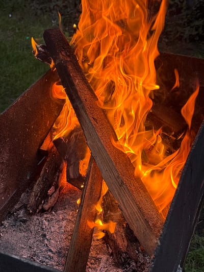 A close-up, overhead shot captures a fire burning within a metal fire pit. The fire pit is rectangular and appears to be made of rusted metal. Inside, several logs are stacked, with a larger log prominently placed diagonally across the frame. The logs are engulfed in vibrant orange and yellow flames, with white smoke rising and swirling. The base of the fire pit is filled with gray ash and embers. The background is blurred, showing green grass and some dark foliage, suggesting an outdoor setting. No people are visible in the image. The scene appears to be located in the Ганчарскі сельскі Савет, Belarus.