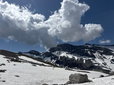 This is a photograph of a snow-covered mountain landscape under a partly cloudy blue sky. The foreground is a gentle slope covered in snow with scattered rocks. Further up the slope, darker, rocky terrain is visible, interspersed with patches of snow. In the background, a rugged mountain range with steep, dark rock faces and significant snow cover dominates the scene. Large, white cumulus clouds are prominent in the sky. The image appears to be taken in a high-altitude alpine environment. The scene is devoid of any people or man-made structures.